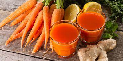2 glasses containing a fresh carrot smoothie. Fresh carrots, lemons and ginger root surround the glasses.