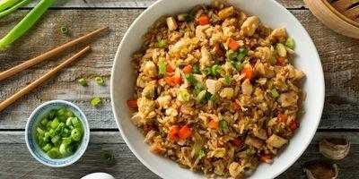 White bowl filled with brown rice, bite-size pieces of chicken, carrots, peas and garnished with slices of green onions.