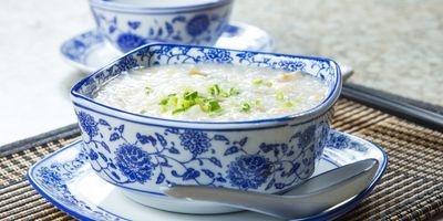 White porcelain bowl with blue flower pattern filled with congee garnished with green onions. The bowl is on a matching saucer with a spoon resting by it.