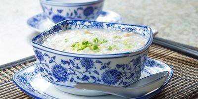 White porcelain bowl with blue flower pattern filled with congee garnished with green onions. The bowl is on a matching saucer with a spoon resting by it.