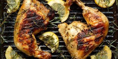 Two charred chicken legs on a grill garnished with lemon slices and fresh rosemary