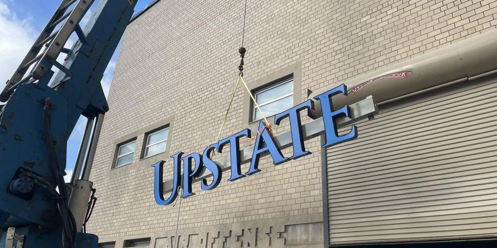 SIGN OF THE TIMES: Crews lift “Upstate” into place on the Onondaga County War Memorial. A sign announcing the official name of the building “the Onondaga County War Memorial Upstate Medical University Arena” was in place by week’s end.
