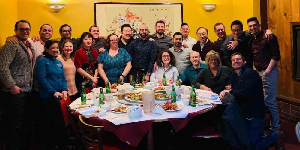 Image of a diverse group of twenty-one adults sitting and standing behind a round table after finishing dinner, posing for a group photo