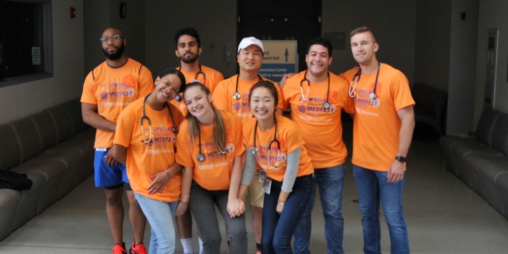 Image of a diverse group of students wearing matching orange t-shirts that say "Special Olympics MedFest," smiling and posing for a group photo indoors
