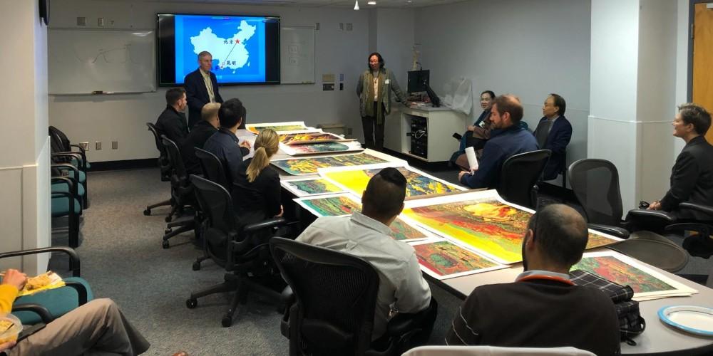 Image of a group of adults sitting around a table in a conference room. The table has colorful artwork on it, and there are two presenters at the front of the room near a projected slide depicting a map of china.