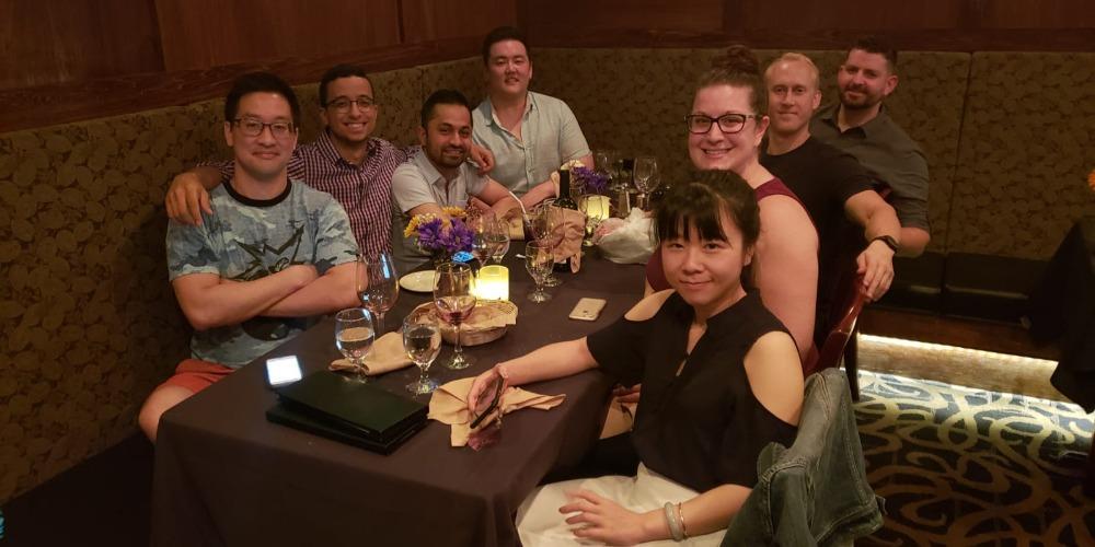 Image of a diverse group of eight people in their twenties, sitting at a booth in a restaurant with glasses of water, turning to smile at the photographer.