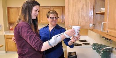 Therapist and patient stand putting away dishes with a Bioness therapy device on the patients left arm.
