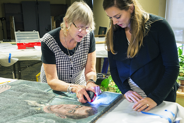 Art conservator Susan Blakney uses an LED light to examine the World War II portrait of Arthur Ecker, MD, PhD, founder of the neurosurgery department at Upstate Medical University. Cara Howe, Upstate's curator of historical collections, observes. (PHOTO BY WILLIAM MUELLER)
