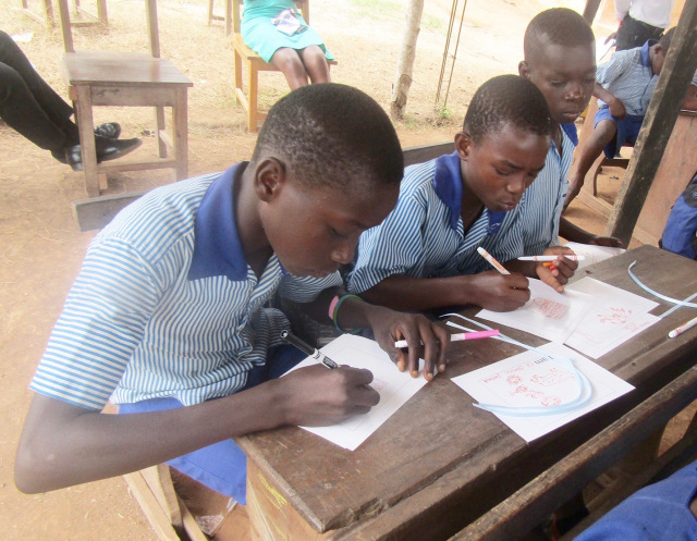 These young men are making their books during the literacy program. They walk four hours to attend school in an outdoor classroom with a dirt floor and metal roof supported by tree branches. (PHOTO BY SUSAN KEETER)