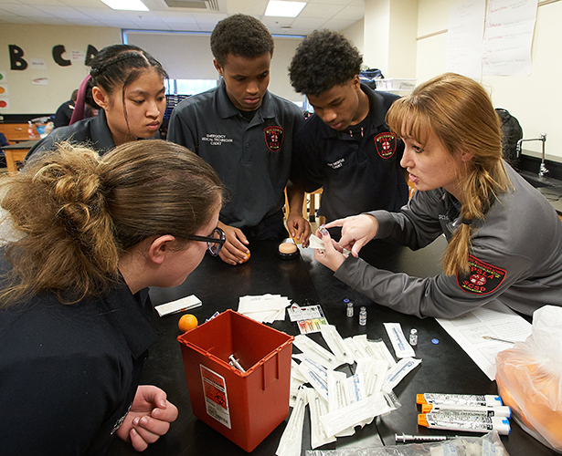 Brandi Schaefer, right, prepares students to give injections, which they practice using oranges. Pictured, clockwise from left, are Alex Jones, Mya Aung, Ridwan Sirad and Hussein Musa.
